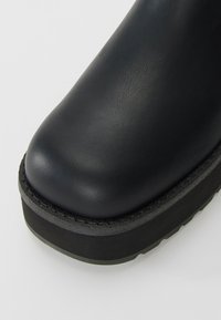 Bout de chaussure en cuir noir avec une semelle épaisse en caoutchouc noir texturé, montrant une surface supérieure lisse et une construction robuste.