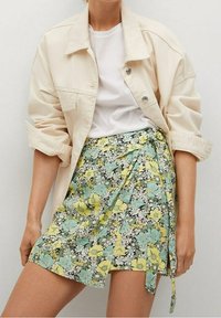 Veste crème sur un t-shirt blanc, associée à une jupe portefeuille à motif floral en vert, jaune et noir. Ourlet court, nouée à la taille.