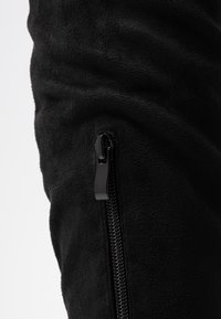 Particolare di un tessuto in pile nero con una cerniera nera verticale parzialmente chiusa su uno sfondo chiaro uniforme.
