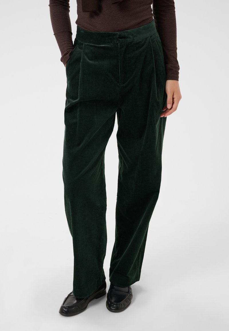 Personne portant un pantalon large en velours côtelé vert foncé avec des mocassins noirs et un haut à manches longues foncé, mains détendues le long du corps.