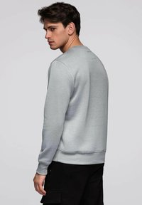 Młody mężczyzna w szarej bluzie i czarnych spodniach stoi bokiem, spoglądając przez ramię na jednolitym tle.