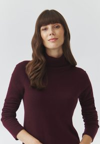 Burgundy színű garbó pulóver, puha anyagból készült, hosszú ujjal és testhezálló fazonnal, bordázott nyakkivágással és sima felülettel.