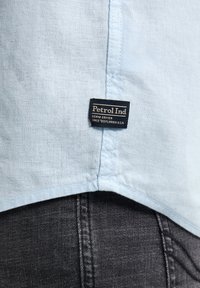 Țesătură de cămașă albastru deschis cu un țesut texturat, având o etichetă de marcă neagră care scrie "Petrol Ind" și blugi gri închis dedesubt.