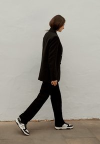 Blazer nero su misura abbinato a pantaloni coordinati, indossato con sneakers nere e bianche. Lo sfondo è un muro chiaro e la figura sta camminando.