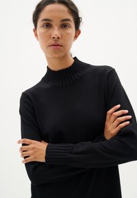 Svart stickad tröja med en räfflad hög krage och räfflade ärmar, som visar upp en slim-fit silhuett och mjuk textur.