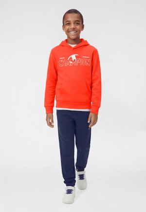 Ragazzo sorridente che cammina in avanti indossando una felpa con cappuccio Champion di un arancione brillante, pantaloni della tuta blu navy e scarpe da ginnastica bianche con lacci blu navy.