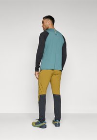 Langærmet skjorte i teal med sorte ærmer, parret med gule og sorte bukser. Har kontrasterende farver og et sporty design.
