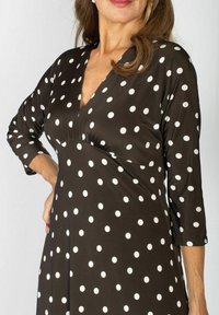 Kobieta w czarnej sukience w białe kropki, z rękawami do trzech kwart, z dekoltem w serek, pozująca z jedną ręką na biodrze.