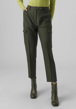 Kobieta ubrana w oliwkowozielone spodnie typu cargo z przednimi i bocznymi kieszeniami, zestawione z pasującymi zielonymi botkami do kostki i żółtym prążkowanym swetrem.