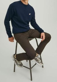 Navyblaues Sweatshirt mit rundem Ausschnitt und kleinem Logo, kombiniert mit braunen Hosen. Tragend mit grauen und weißen Sneakers, sitzt auf einem Hocker.