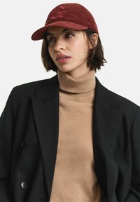 Rote Cord-Kappe mit einem gestickten "G", kombiniert mit einem beigen Rollkragenpullover und einem schwarzen Blazer, die einen geschichteten, strukturierten Look zeigen.