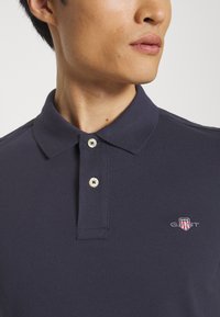 Mann trägt ein dunkelblaues Poloshirt mit zwei geschlossenen Knöpfen und einem kleinen bestickten GANT-Logo in Rot, Weiß und Blau auf der Brust.