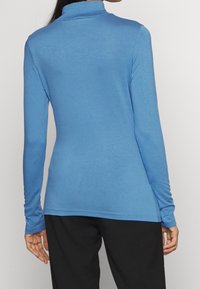 Långärmad, figurnära turtleneck i ljusblått tyg. Har en slät textur och en rak nederkant. Bärs med svarta byxor.