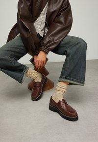 Bruine leren schoenen met witte stiksels, gelaagde zool en ronde neus, gecombineerd met beige geribbelde sokken, gedragen met losse blauwe jeans.