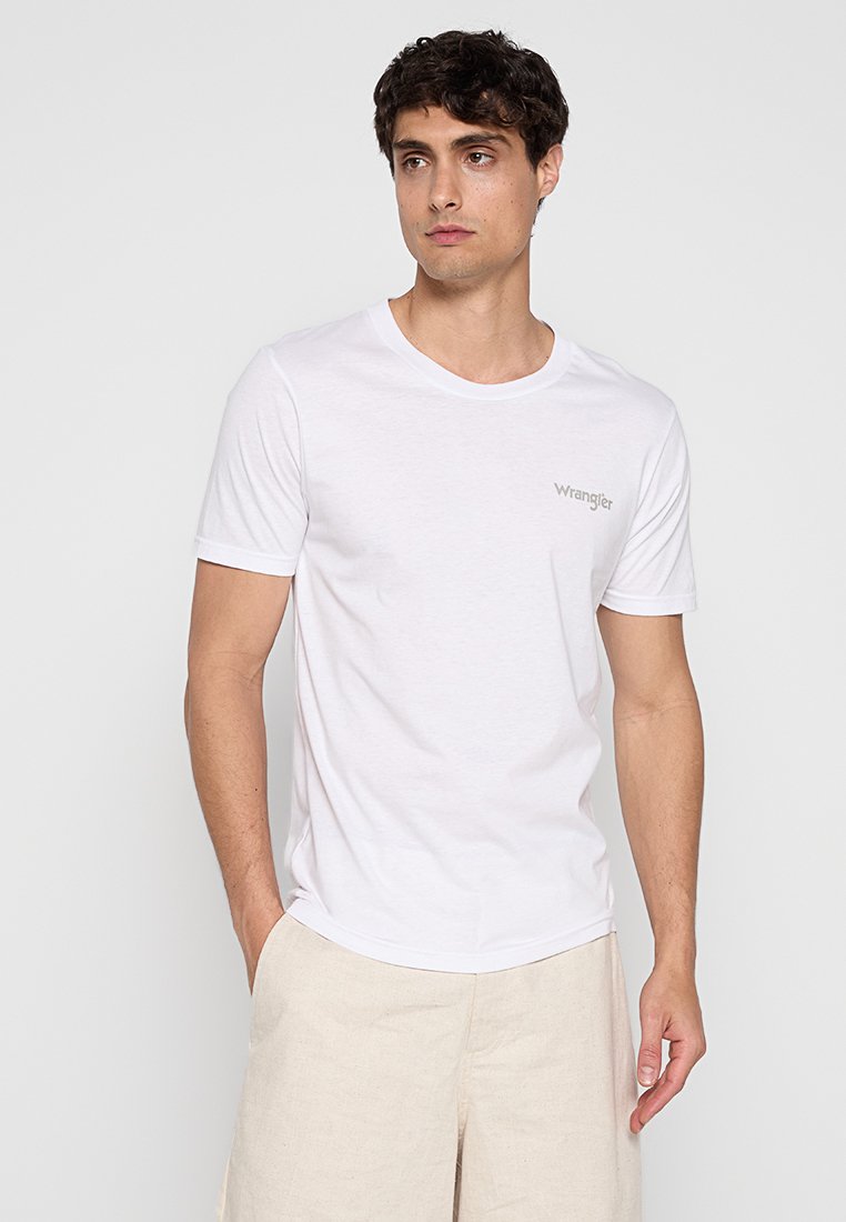 Mężczyzna w białej koszulce Wrangler i beżowych szortach, stojący z jedną ręką w kieszeni na tle jasnego, jednolitego tła.