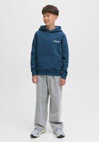 Felpa blu scuro realizzata in tessuto morbido, con tasca frontale e logo discreto. Abbinata a jeans larghi grigio chiaro e sneakers.
