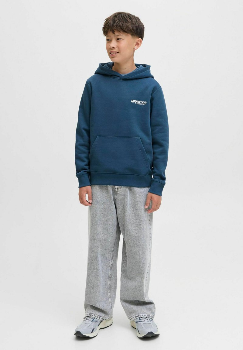 Felpa blu scuro realizzata in tessuto morbido, con tasca frontale e logo discreto. Abbinata a jeans larghi grigio chiaro e sneakers.