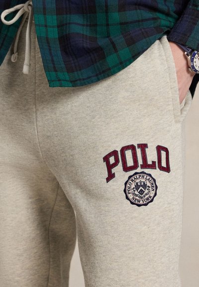 Άτομο που φοράει μπεζ φόρμα με "POLO" και λογότυπο στο μηρό, πράσινο και ναυτικό πουκάμισο σε καρό σχέδιο και ένα ρολόι χειρός με μπλε λουρί.