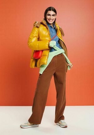 Gul pufferjakke, blå denimskjorte, mintgrøn sweater, brune bukser, rød crossbody taske og lyse sneakers mod en orange baggrund.