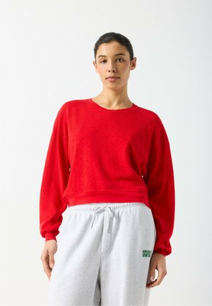 Maglione rosso con texture, maniche lunghe e scollo rotondo, abbinato a pantaloni della tuta grigi con un logo verde sul lato.