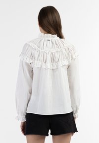 Camicia bianca con volant, collo alto, tessuto testurizzato, maniche lunghe a sbuffo e dettagli arricciati sul retro. Abbinata a pantaloni corti neri.
