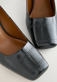 Czarne, patentowane skórzane buty o kwadratowym nosku z tłoczonym wzorem imitującym skórę krokodyla i gładkim, brązowym wnętrzem. Minimalistyczna konstrukcja z niskim obcasem.