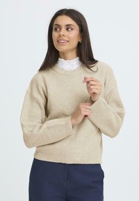 Bežový rebrovaný sveter s okrúhlym výstrihom a voľným strihom, má dlhé rukávy a skrátený lem. Nosený cez bielu košeľu s golierom.