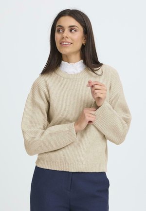 Beige ribbad tröja med rund halsringning och lös passform, med långa ärmar och kortad nederkant. Buren över en vit skjorta med krage.