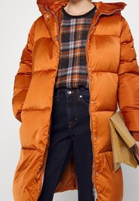 Vadderad orange kappa med huva och tryckknappsstängning, tillsammans med en rutig tröja och mörka jeans, håller en brun papperspåse.