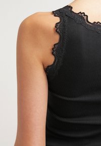 Sort ribbet tank top med bølgemønstrede blonder langs halsudskæringen og stropperne, der fremhæver en tætsiddende silhuet og blød tekstur.