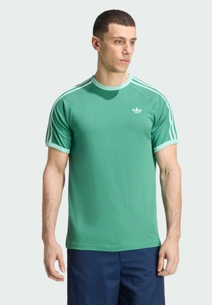 Homme portant un t-shirt Adidas vert avec des rayures vert clair sur les manches et un logo, associé à un pantalon bleu foncé, debout devant un fond uni.