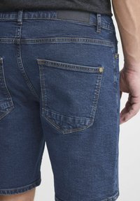 Tamne traperice u tamnoplavoj boji s glatkom teksturom, koje imaju dva stražnja džepa i kontrastne šavove. Minimalistički detalj crnog struka.