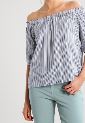 Persoon draagt een blauw-wit gestreepte off-shoulder blouse met driekwart mouwen en lichtblauwe broek, hand rust op heup.