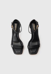 Para czarnych skórzanych sandałów Michael Kors z kwadratowym noskiem, paskiem wokół kostki i niskim obcasem na białym tle.