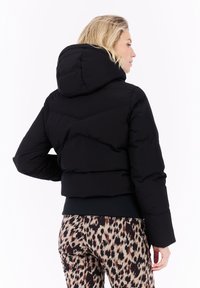 Mujer con cabello rubio, vestida con una chaqueta acolchada negra con capucha y pantalones de estampado de leopardo, de espaldas contra un fondo blanco.