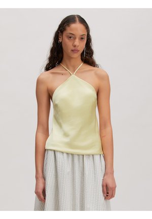 Femme aux longs cheveux bouclés portant un haut dos-nu jaune pâle et une jupe légère rayée, debout devant un fond uni.