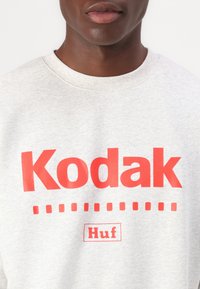 Γκρι φούτερ από βαμβάκι, με μεγάλο κόκκινο κείμενο "Kodak" και λογότυπο "Huf" από κάτω, με μοτίβο τελείας κάτω από την επωνυμία.