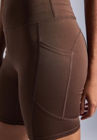 Pantaloni scurți de sport marron, confecați dintr-un material elastic, având cusături plate și o buzunar lateral. Designul este ajustat, cu o talie medie.
