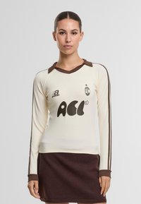 Pitkähiha beige paita, jossa on ruskea kaulus ja hihansuut, sisältäen graafisen logon ja raidat hihojen varrella. Yhdistetty tummanruskeaan hameeseen.