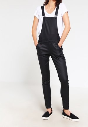 Kvinde iført sorte tætsiddende overalls med justerbare stropper, en hvid t-shirt og sorte slip-on sko, stående med hænderne i lommerne.