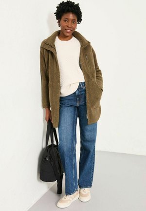 Olivgrüne Fleecejacke mit Druckknöpfen, cremefarbener strukturierter Pullover, blaue Jeans mit geradem Bein und beiges Sneaker, hält eine schwarze Tasche.