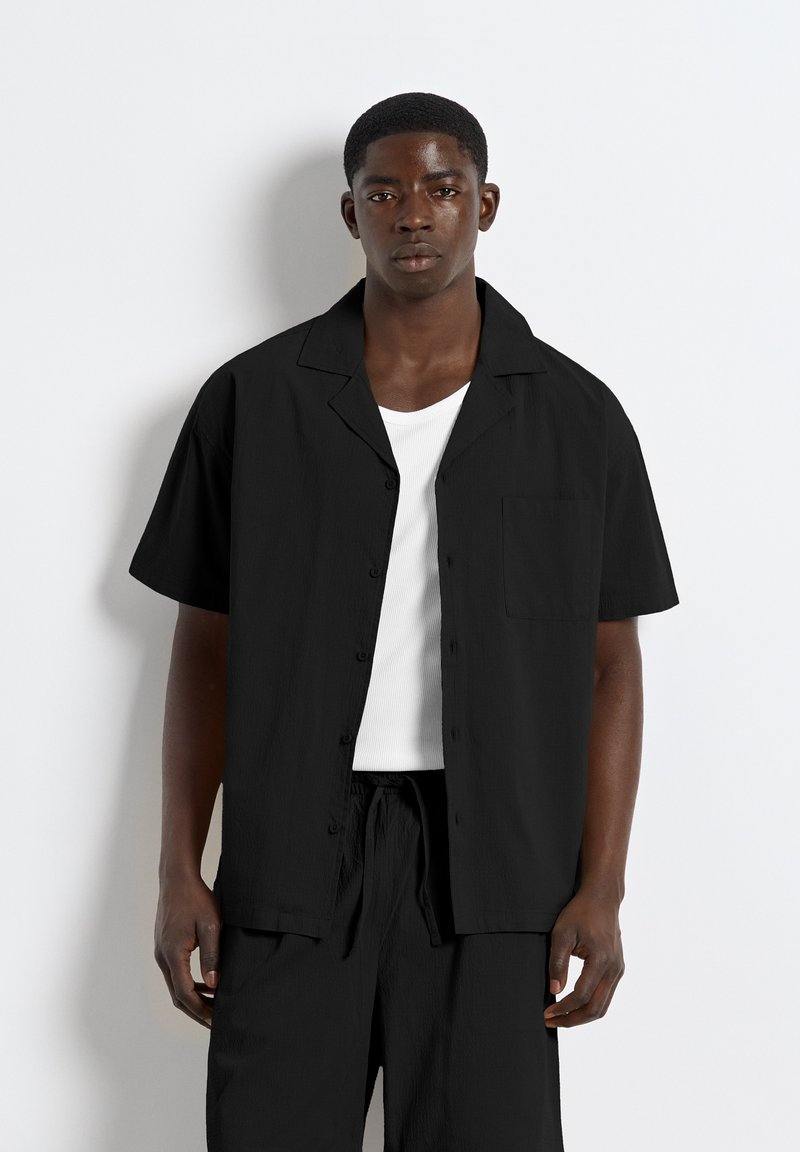 Jeune homme debout portant une chemise noire à manches courtes boutonnée sur un débardeur blanc avec un pantalon noir sur un fond clair uni.