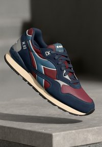 Blue and burgundy sneaker with light blue accents, featuring a textured upper and laces, suspended above a stone surface.