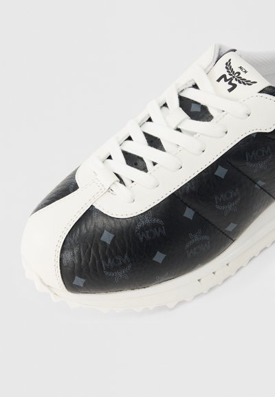 Μαύρο δερμάτινο αθλητικό παπούτσι με λευκές λεπτομέρειες, διαθέτοντας υφή στην επ ιφάνεια, κορδόνια στο μπροστινό μέρος και τυπωμένα λογότυπα σε όλο το παπούτσι. Επίστρωση από καουτσούκ με αντιολισθητική σόλα.