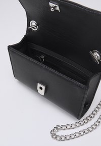 Sac bandoulière en cuir noir avec une finition texturée, une bandoulière en chaîne argentée et une fermeture à bouton-pression. L'intérieur comprend des emplacements pour cartes et une poche zippée.