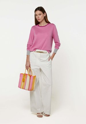 Femme portant un haut à manches longues rose, un pantalon blanc à jambes larges, tenant un sac à main tissé rayé rose, jaune et blanc, debout devant un fond blanc.