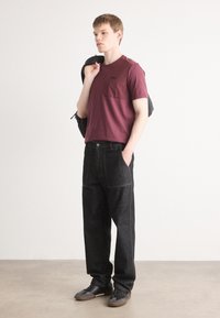 Bordo īsām piedurknēm krekls ar krūšu kabatu, saskaņots ar tumšiem džinsu biksēm. Modelis pozē ar melnu mugursomu uz viena pleca.