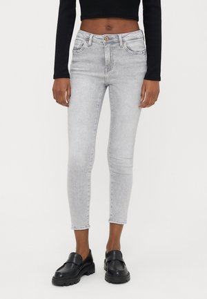 Kvinde iført lysegrå skinny jeans, sort cropped langærmet top og sorte chunky loafers, stående mod en ensfarvet baggrund.