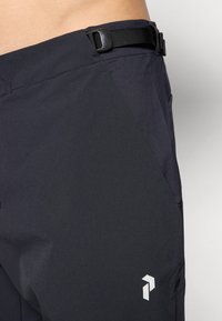 Pantalon de sport noir avec une ceinture, présentant une finition lisse et texturée ainsi qu'un logo blanc bien visible sur le côté. Comprend des poches latérales.
