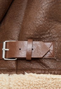 Materiale in pelle marrone con una finitura texturizzata, caratterizzato da una tracolla marrone con una fibbia argentata e dettagli cuciti sui bordi.